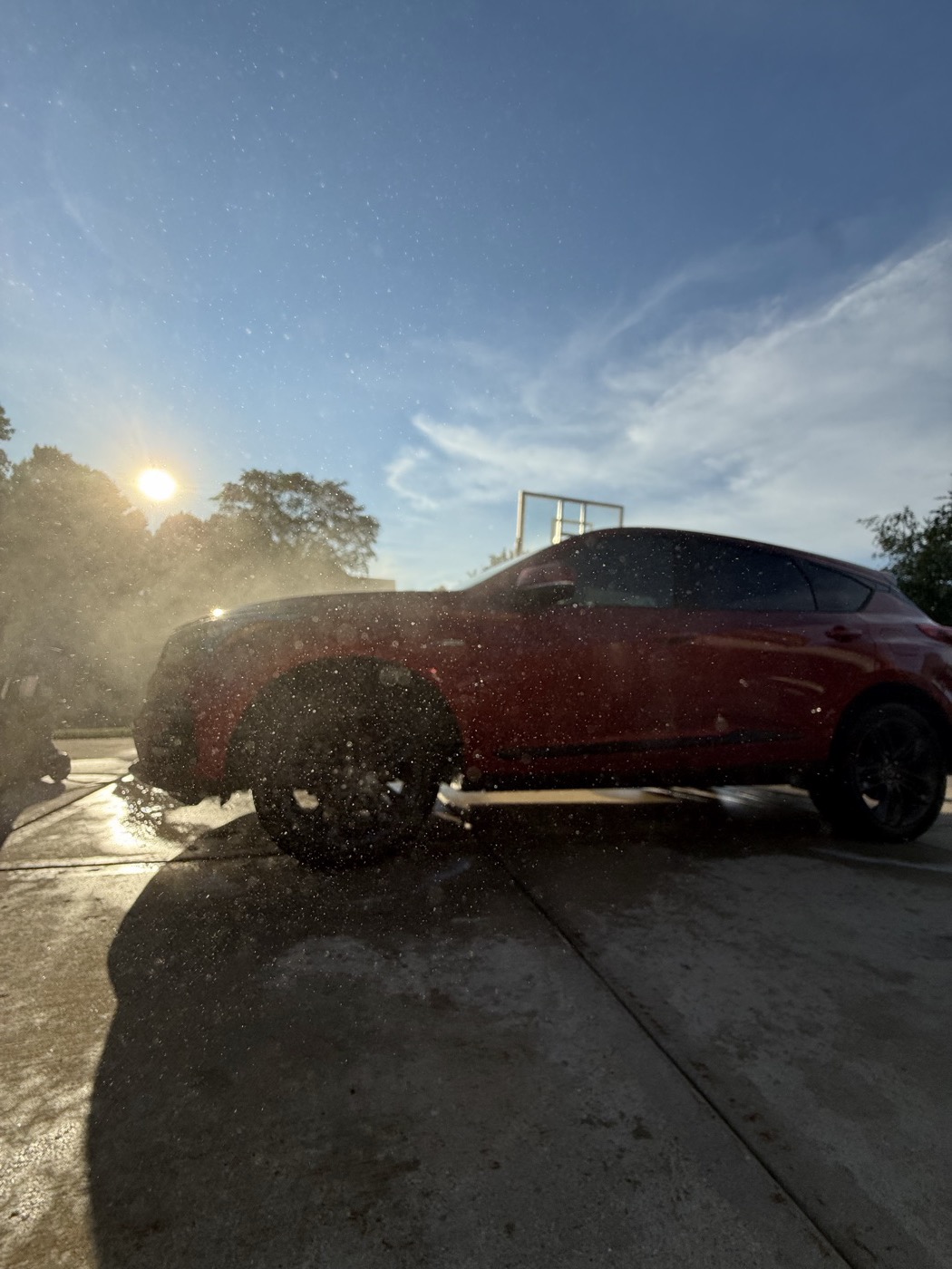 Red Acura low-angle side profile during detail, water spray catching the sun